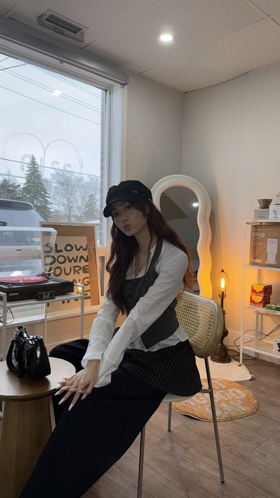 Kaitlyn Duan seated by the window of a record and coffee shop, wearing a black cap and vest