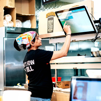 A kitchen staff member reaching up to read and tap the display mounted above their station
